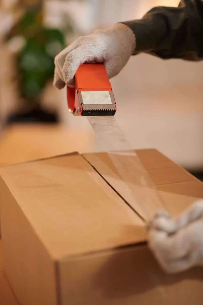 Primer plano de una mano sellando una caja de cartón con cinta de embalaje transparente.