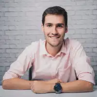 Hombre joven con camisa blanca y reloj, sonriendo frente a un fondo de ladrillo gris.