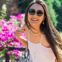 Mujer con gafas de sol, vestida de blanco, posando con una sonrisa frente a flores de color fucsia.