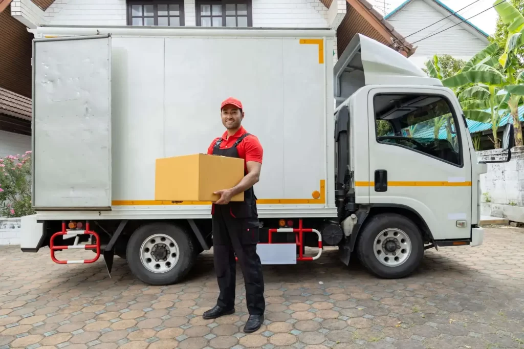 Fotografía de un trabajador de mudanzas con uniforme rojo sosteniendo una caja de cartón frente a un camión de mudanza.
